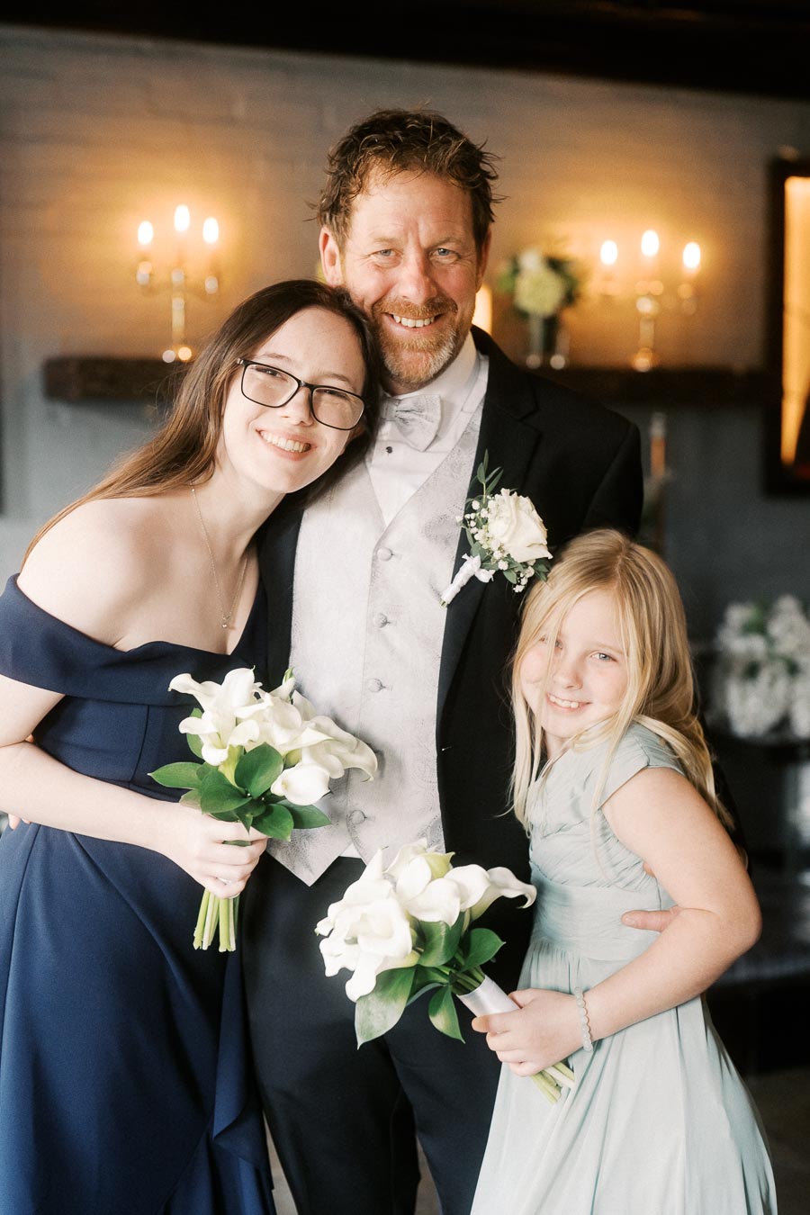 A joyful wedding group portrait featuring a smiling man in a suit and tie alongside two young girls, one wearing glasses and a navy dress, the other in a light blue dress, holding bouquets of white flowers in an elegantly decorated venue with lit candles.