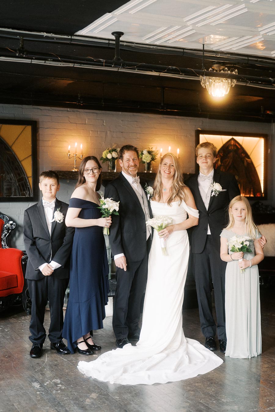 Elegant family portrait at a wedding, featuring the newlywed couple in formal attire surrounded by children, standing indoors with soft lighting and a decorative background.