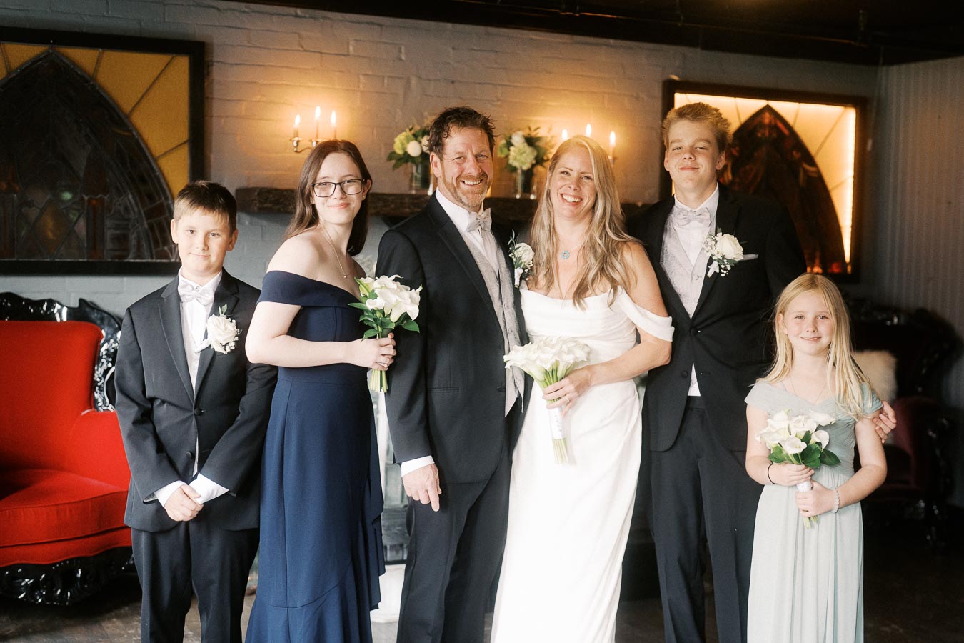 A wedding group portrait featuring a bride in a white dress and the groom in a tuxedo, surrounded by children in formal attire, holding bouquets, in an elegantly decorated room with candles and flowers.