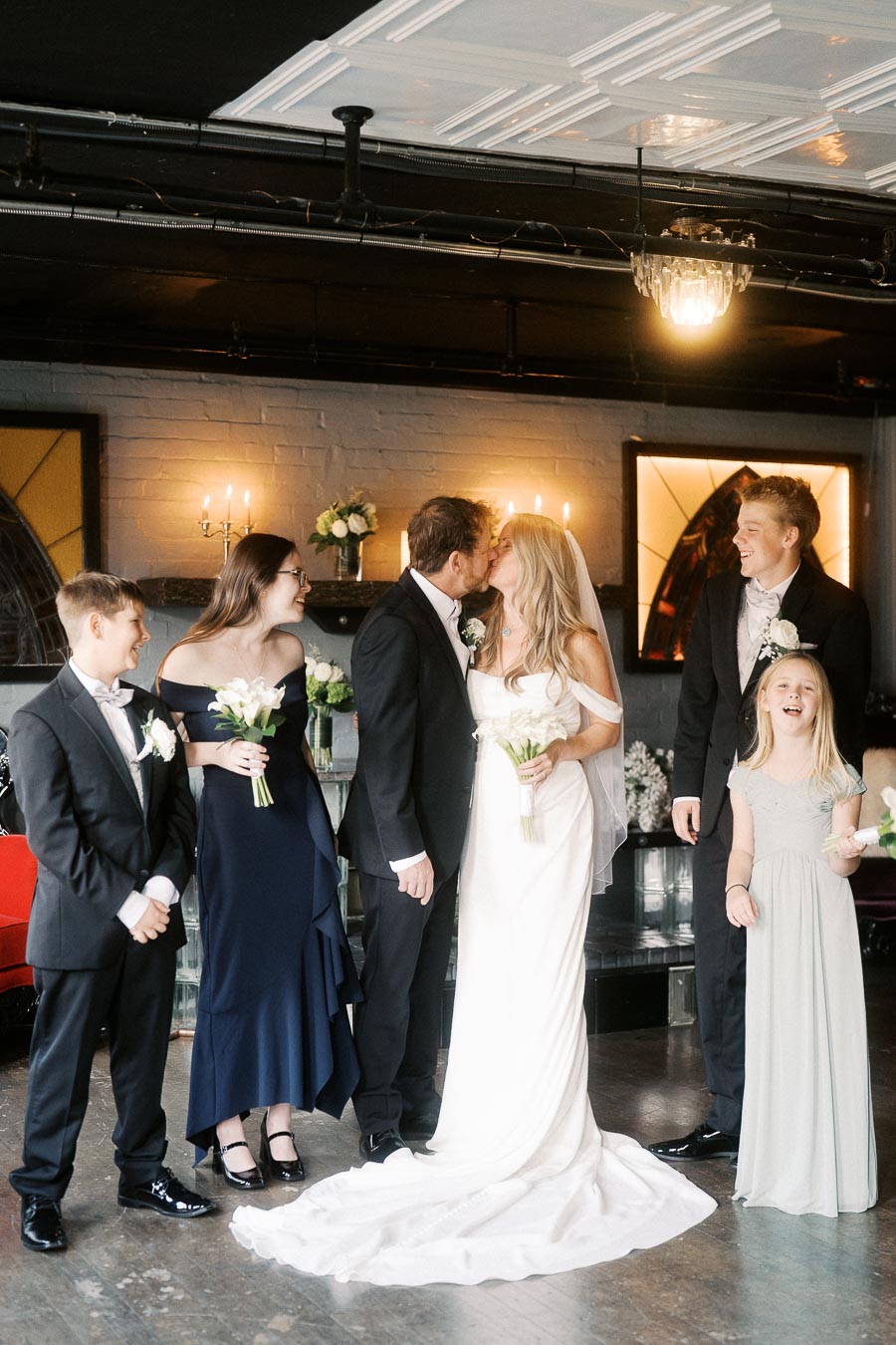 Bride and groom kissing in an elegant indoor wedding ceremony with family, dressed in formal attire with flowers, under soft lighting.