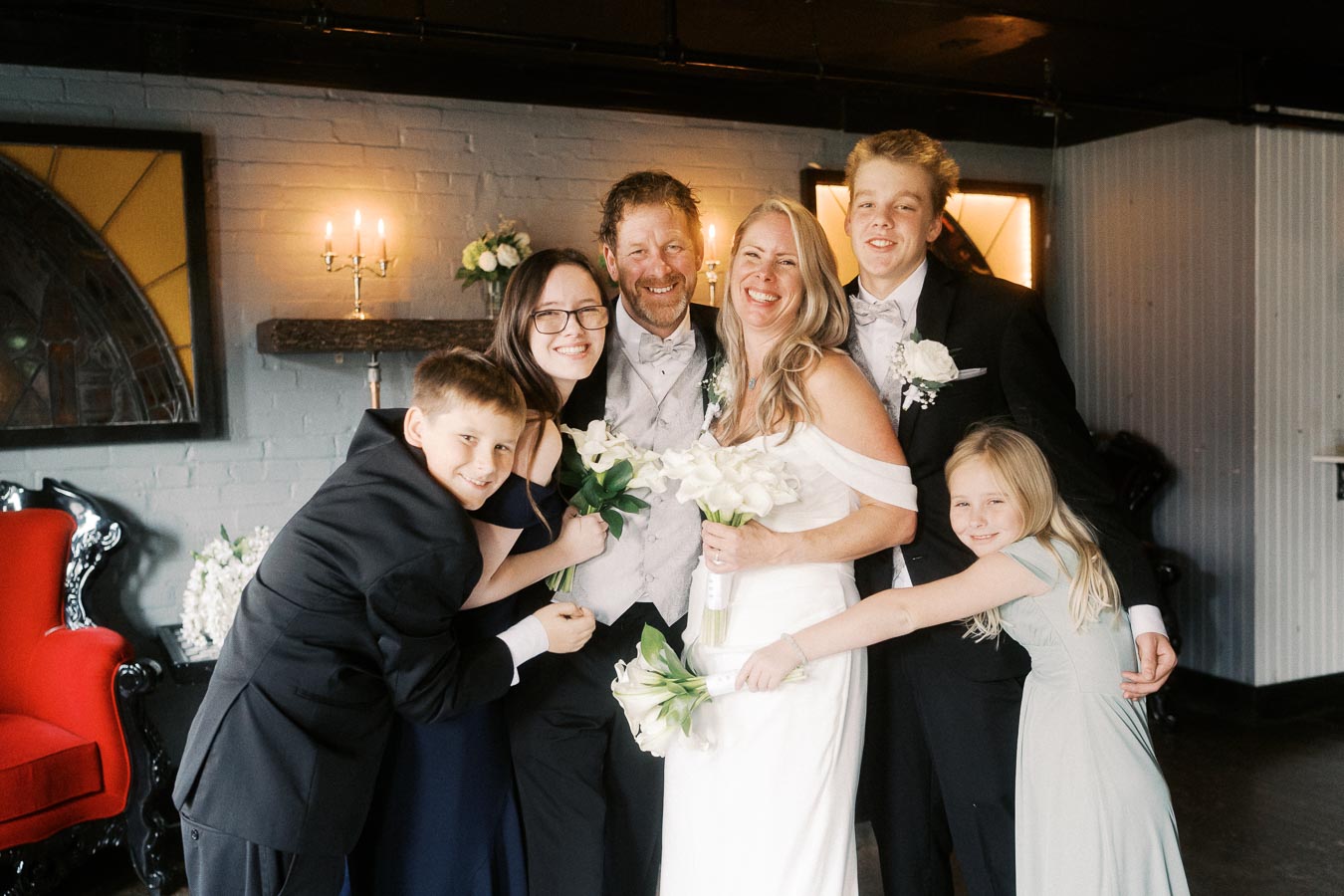 Family posing together at a wedding celebration, featuring the bride in a white dress holding a bouquet, surrounded by children and a beaming groom, in a warmly lit room with elegant decor.