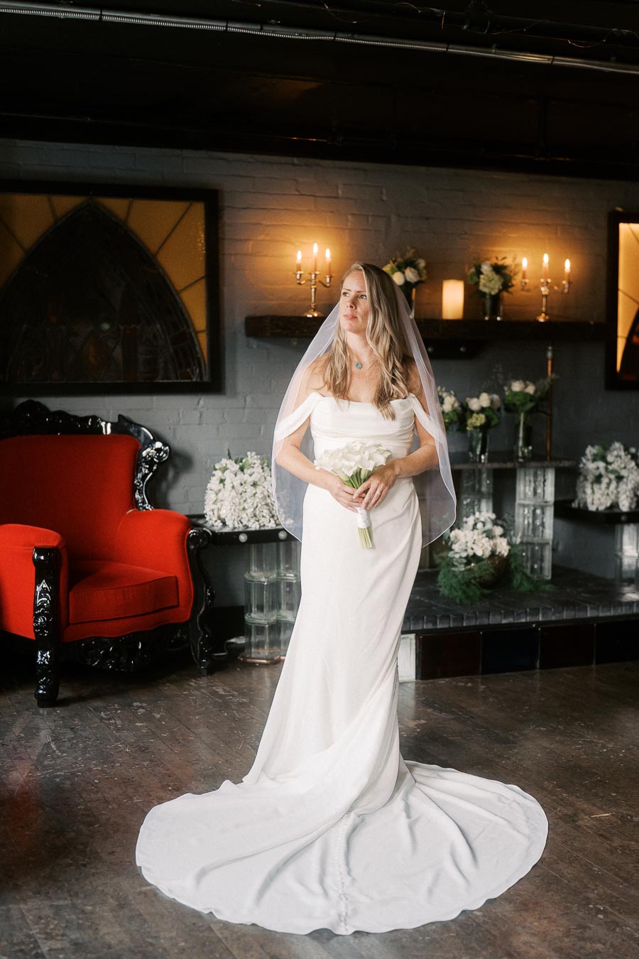 Elegant bride in a flowing white gown holding a bouquet, standing in a beautifully decorated room with a red chair and candlelit ambiance.