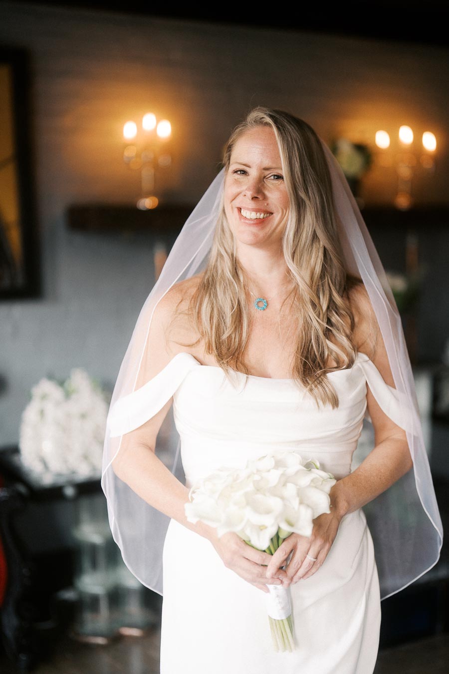 Bride in a white wedding gown holding a bouquet of white flowers, smiling joyfully indoors with soft lighting in the background.