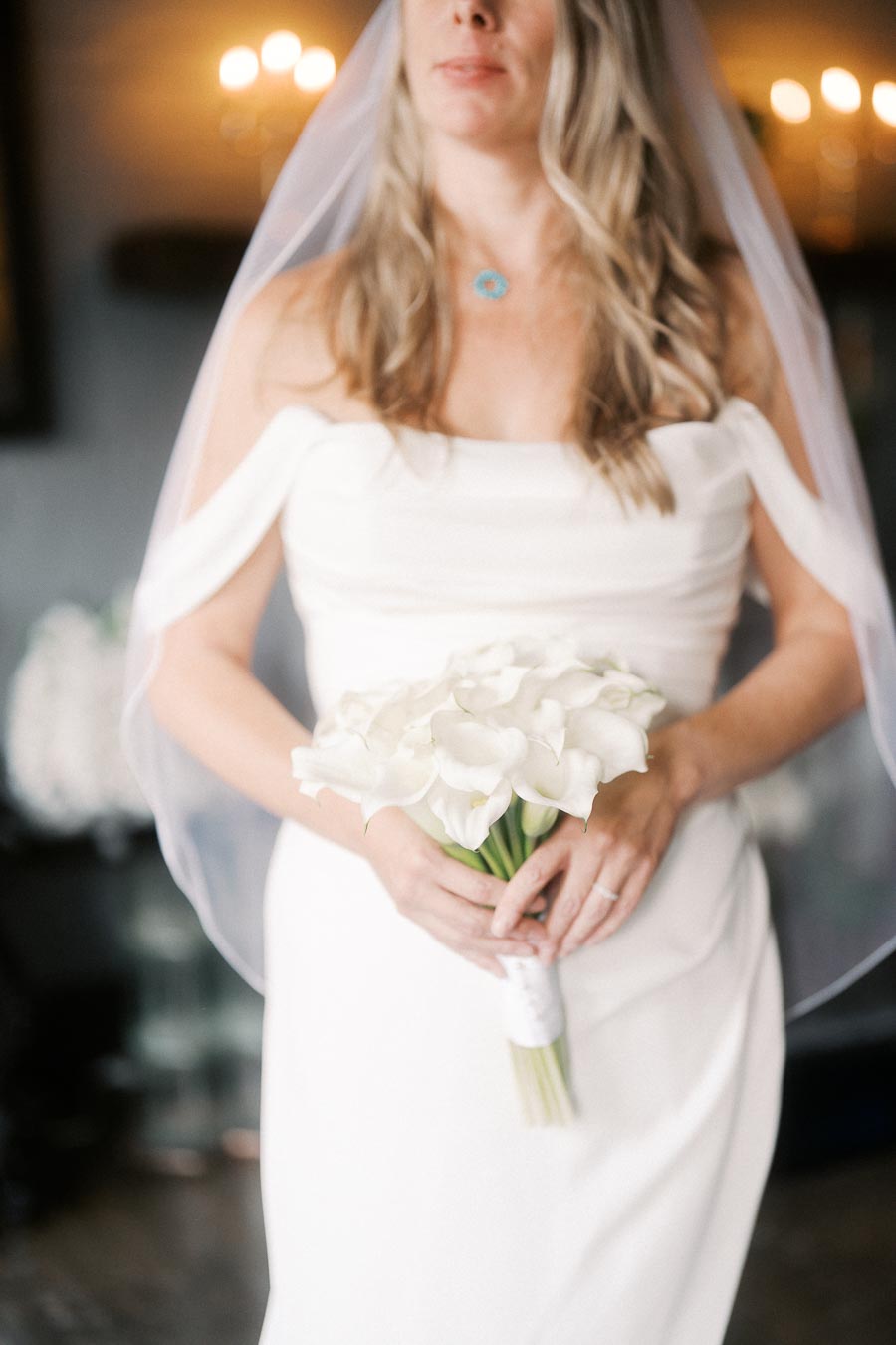 Bride in an elegant white wedding dress holding a bouquet of white calla lilies, wearing a veil and a turquoise necklace, in a softly lit room.