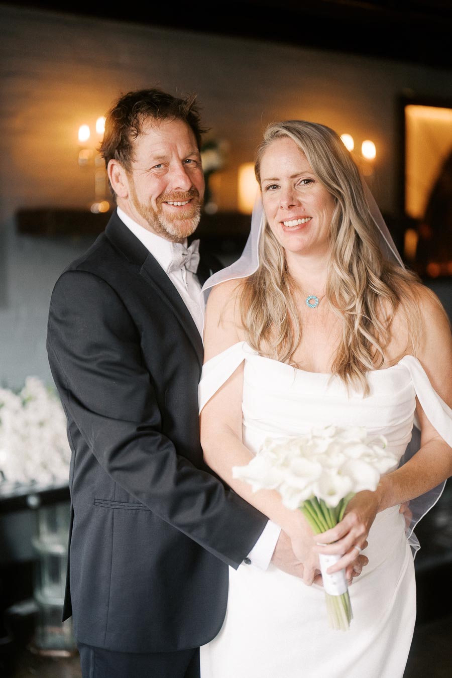 A happy couple dressed in wedding attire, with the groom in a black suit and the bride in a white dress holding a bouquet of white flowers, posing together in a warmly lit room.