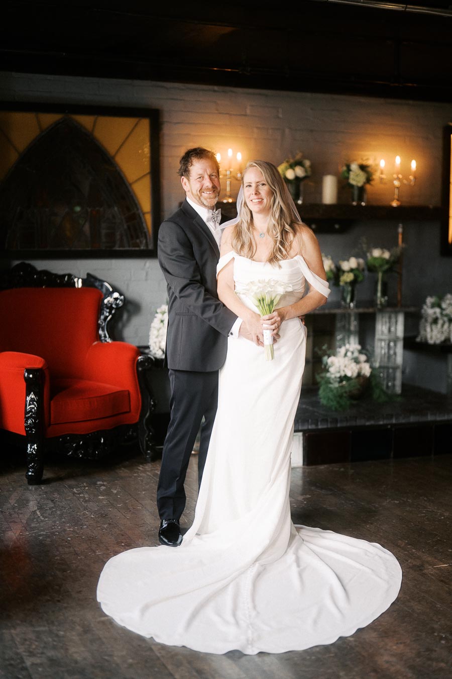 A smiling bride and groom pose indoors in an elegant wedding venue, the bride holding a bouquet of white flowers and wearing a flowing white gown, while the groom is dressed in a black suit. The setting features a red chair and floral decorations.