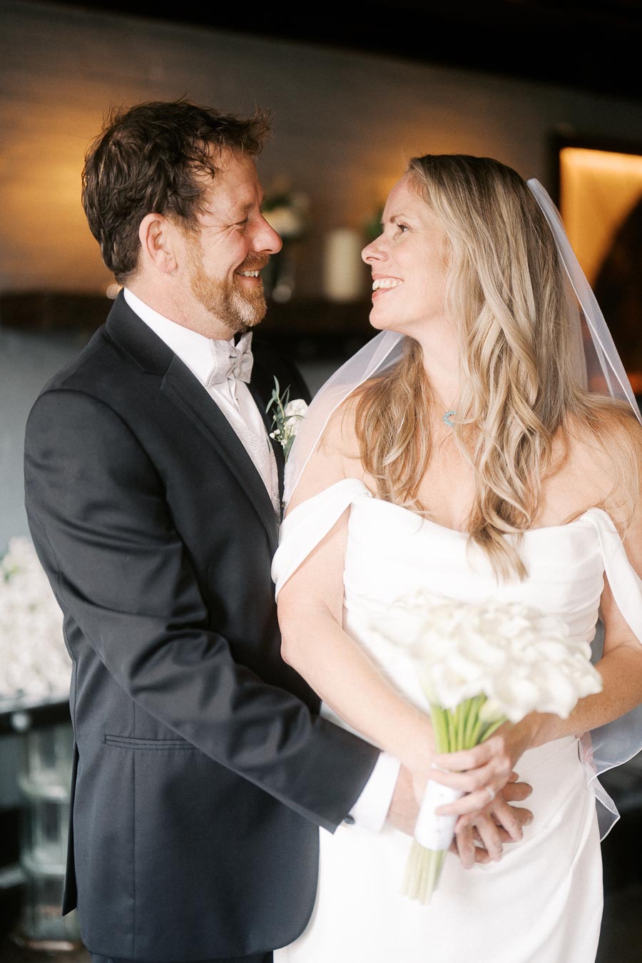 A bride and groom smiling at each other on their wedding day, the groom in a black suit and the bride wearing a white dress and holding a bouquet of white flowers, set in an elegant indoor venue.