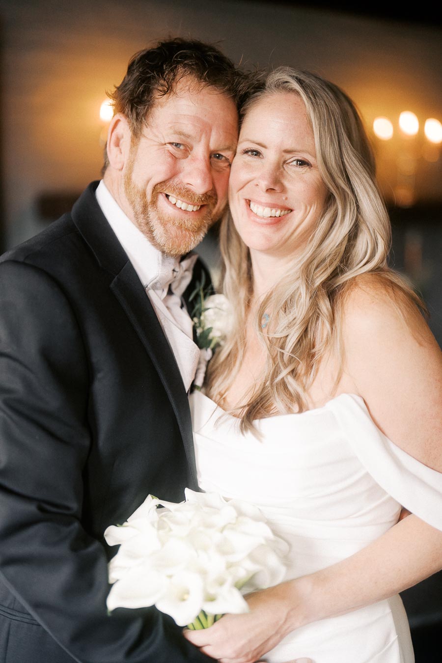 Happy couple on their wedding day, bride in an elegant off-the-shoulder gown holding a bouquet of white flowers, standing next to smiling groom in a black suit. Romantic moment captured indoors with warm lighting.