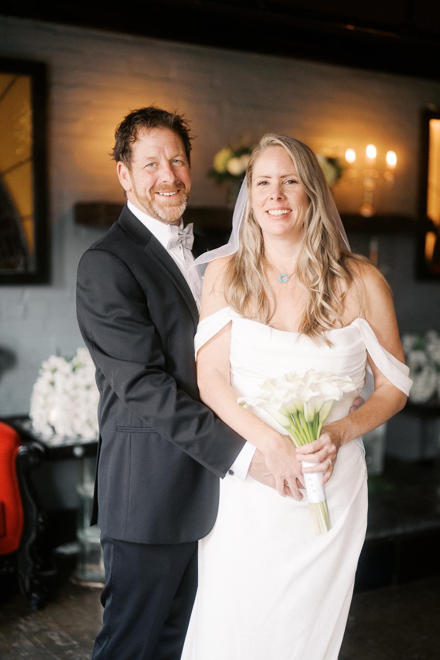 A happy couple in wedding attire, with the groom in a black suit and the bride holding a bouquet, standing in a warmly lit room decorated with flowers and candles.