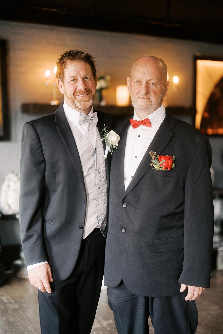 Two men in formal suits with boutonnières, standing together in an elegantly lit room, with candles and soft lighting in the background.