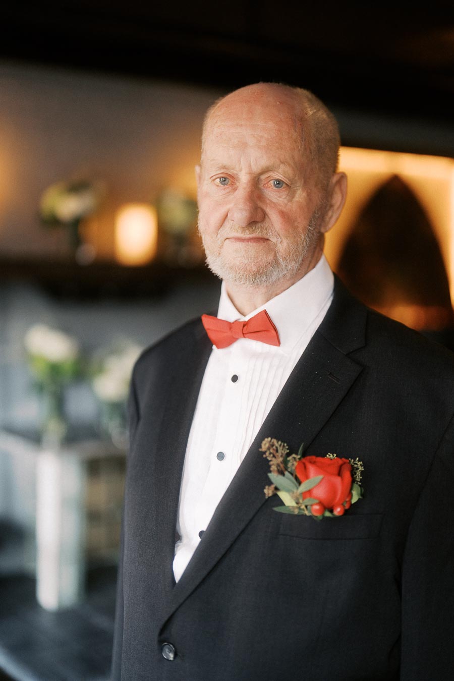Elderly man in formal attire with red bow tie and boutonniere, standing indoors with soft lighting and floral decorations in the background.