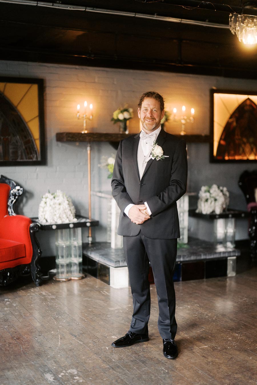 Man in a tuxedo standing in an elegantly decorated room with soft lighting and floral arrangements.