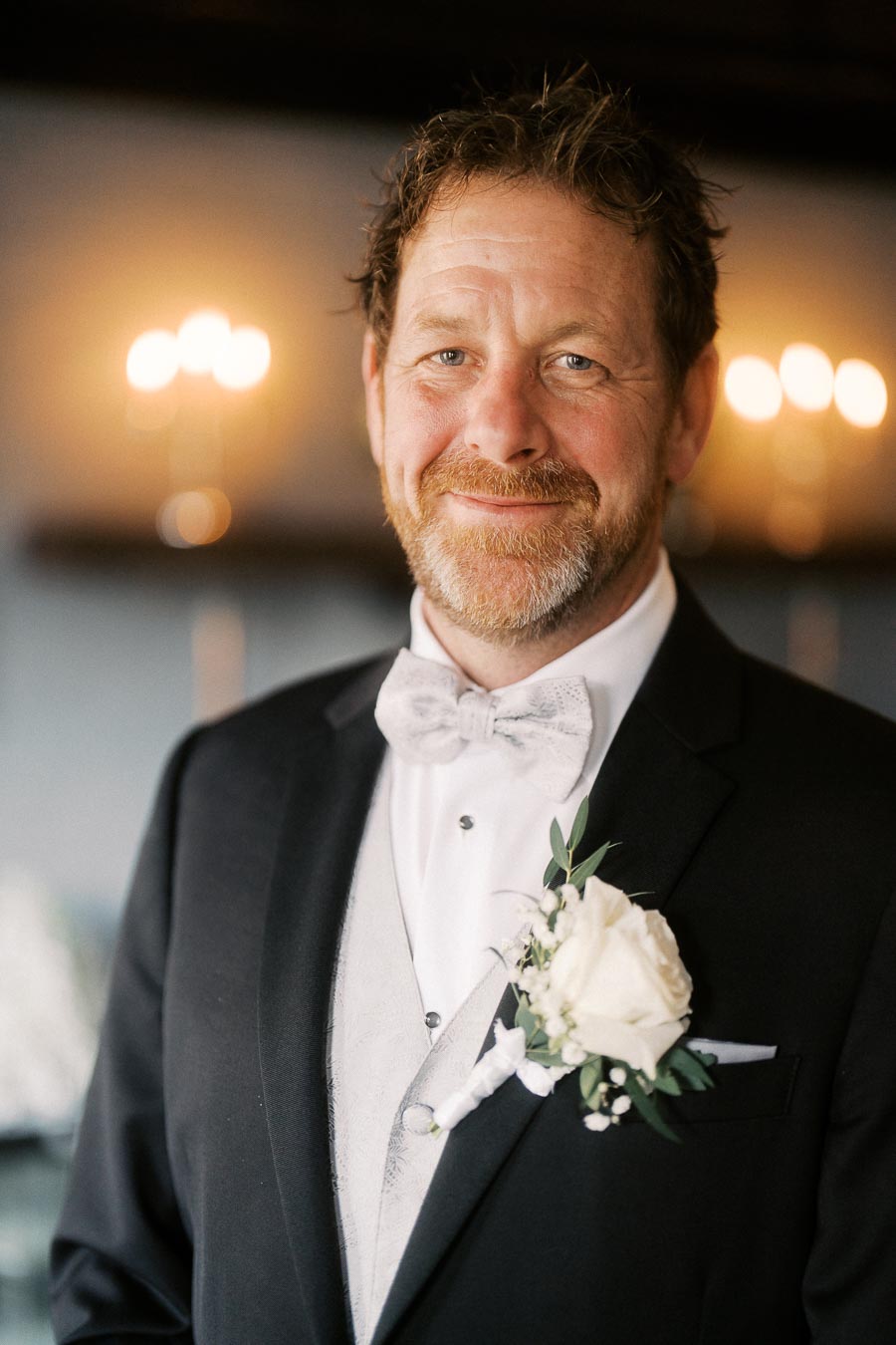 Smiling man in formal attire with a white rose boutonnière, standing in a softly lit setting.