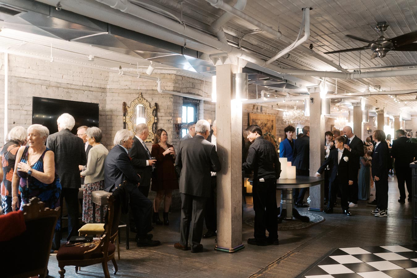 A large group of people socializing at an indoor event, featuring a mix of older and younger individuals in formal attire. The setting has exposed brick walls, elegant decor, and warm lighting, suggesting a sophisticated, celebratory atmosphere.