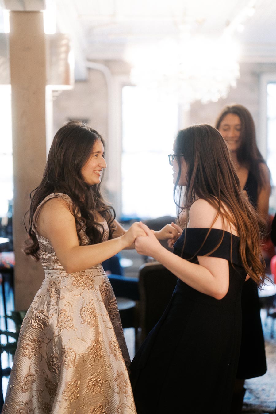 Young women in elegant dresses smiling and holding hands at a social gathering in a brightly lit room.
