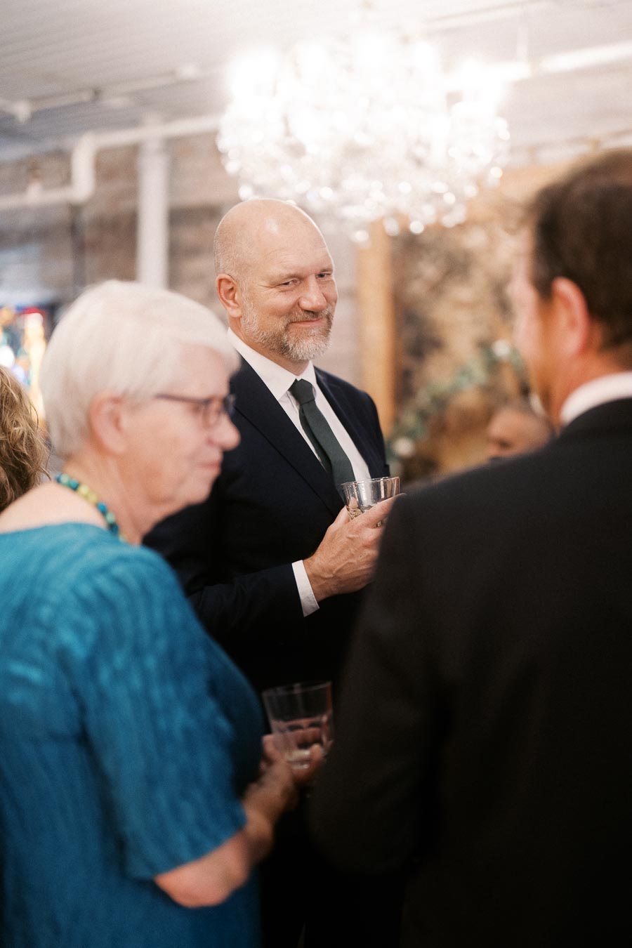 Man in a suit holding a glass, engaged in conversation at an elegant social event with people in formal attire.