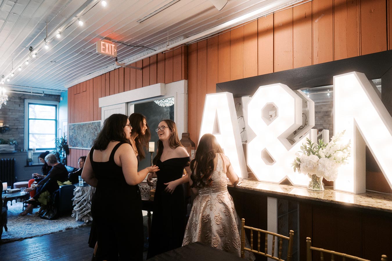 Group of young women in formal attire socializing at an indoor event with illuminated A&M letters in the background.