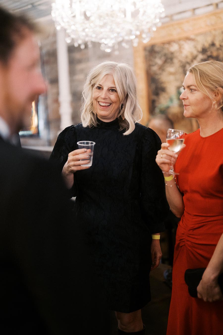 A joyful social gathering with smiling individuals holding drinks, under an elegant chandelier in a warmly lit room.