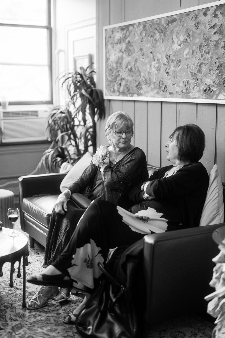 Black and white photo of two women sitting on a sofa in a room with paintings and plants, engaging in conversation. A glass of wine is on a nearby table.