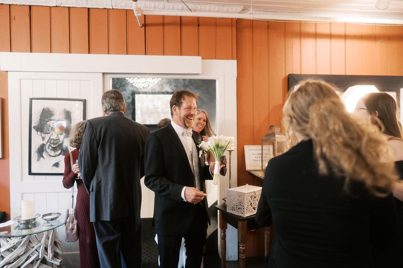 A group of people dressed in formal attire socializing at an indoor event, with one person holding a bouquet of white flowers in a warmly lit room with orange and white walls.