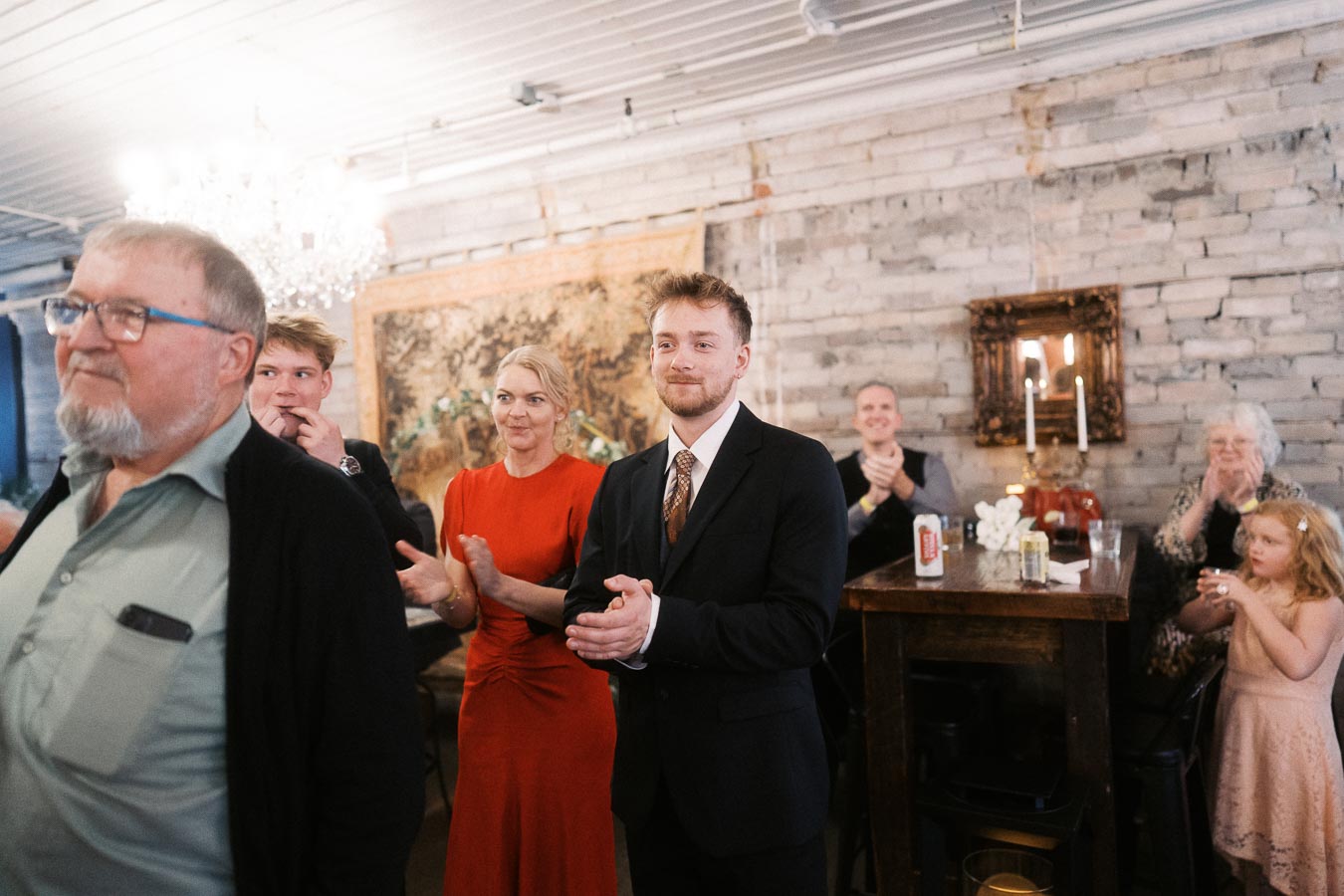A group of people clapping in a warmly lit room with exposed brick walls, featuring a chandelier and decorative artwork in the background. A man in a suit stands prominently in the foreground.