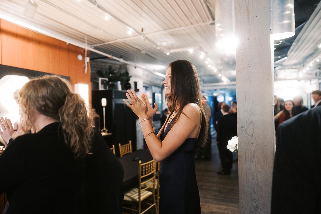 A woman in a navy blue dress clapping and smiling at a formal event held in a decorated indoor venue.