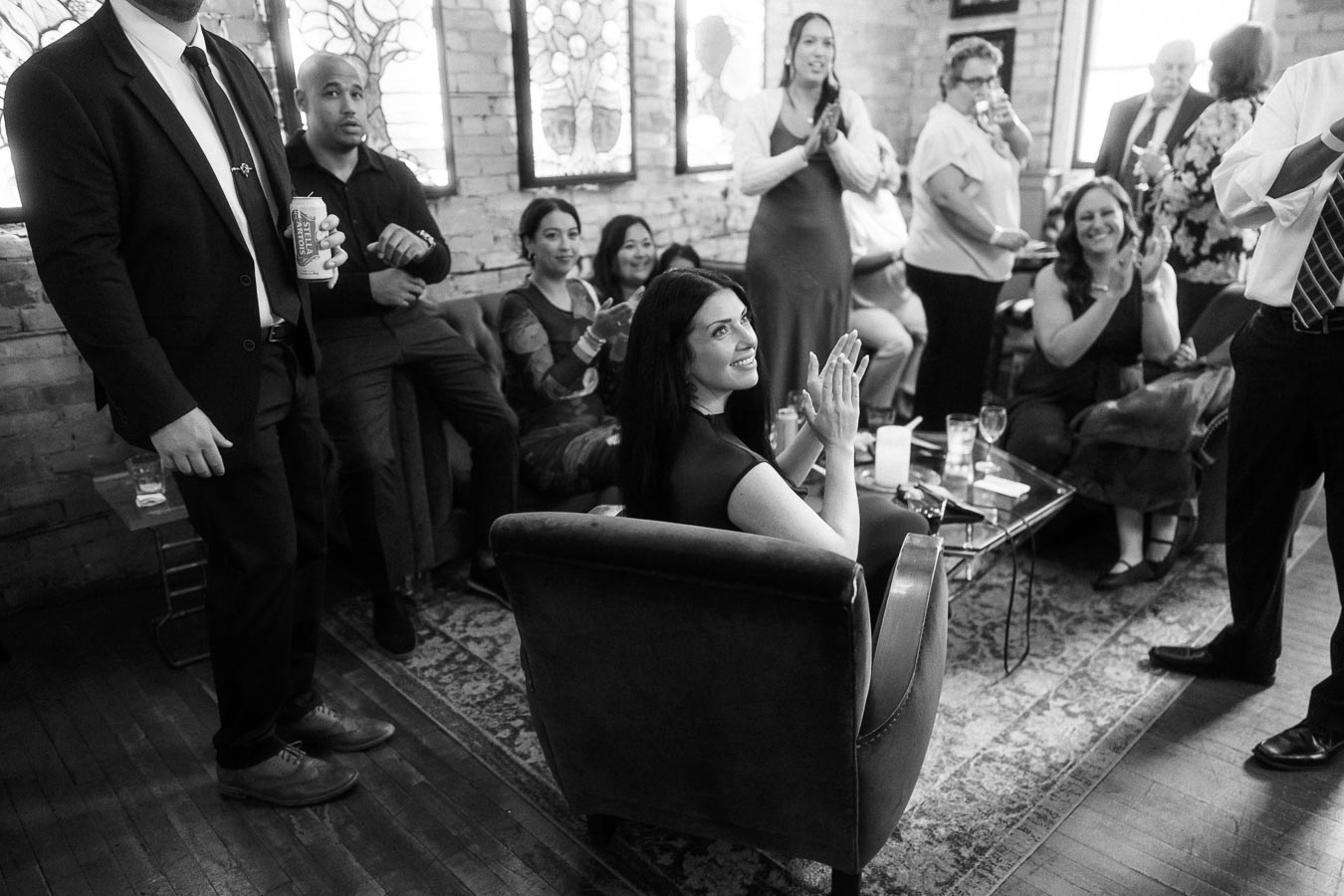 Black and white photo of people clapping and celebrating in a stylish lounge. A woman in the foreground sits in a chair, smiling and applauding, surrounded by a diverse group of casually dressed and business-attired individuals in a warmly lit room with brick walls and large windows.