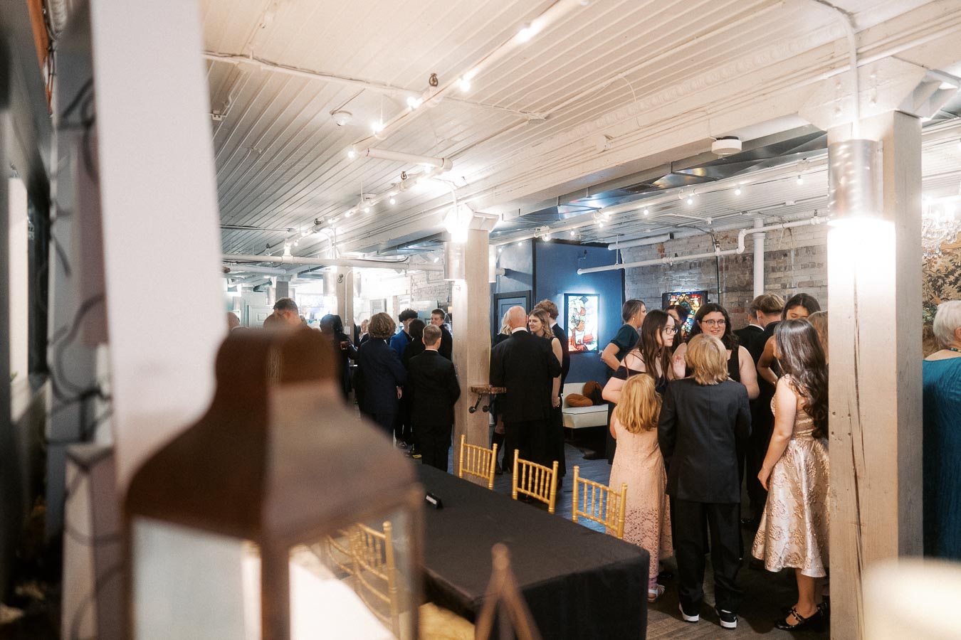 Elegant indoor event with guests in formal attire mingling under string lights in a stylish venue with brick walls.