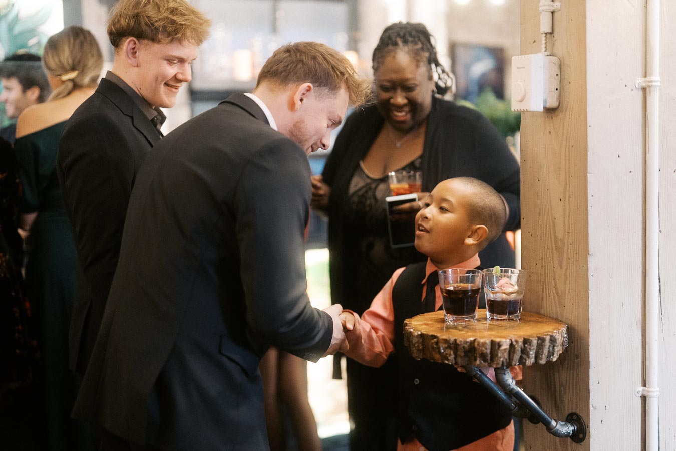A group of people socializing at an indoor event, with a smiling man in a suit shaking hands with a young boy wearing a pink shirt and vest, while others look on with drinks in hand.