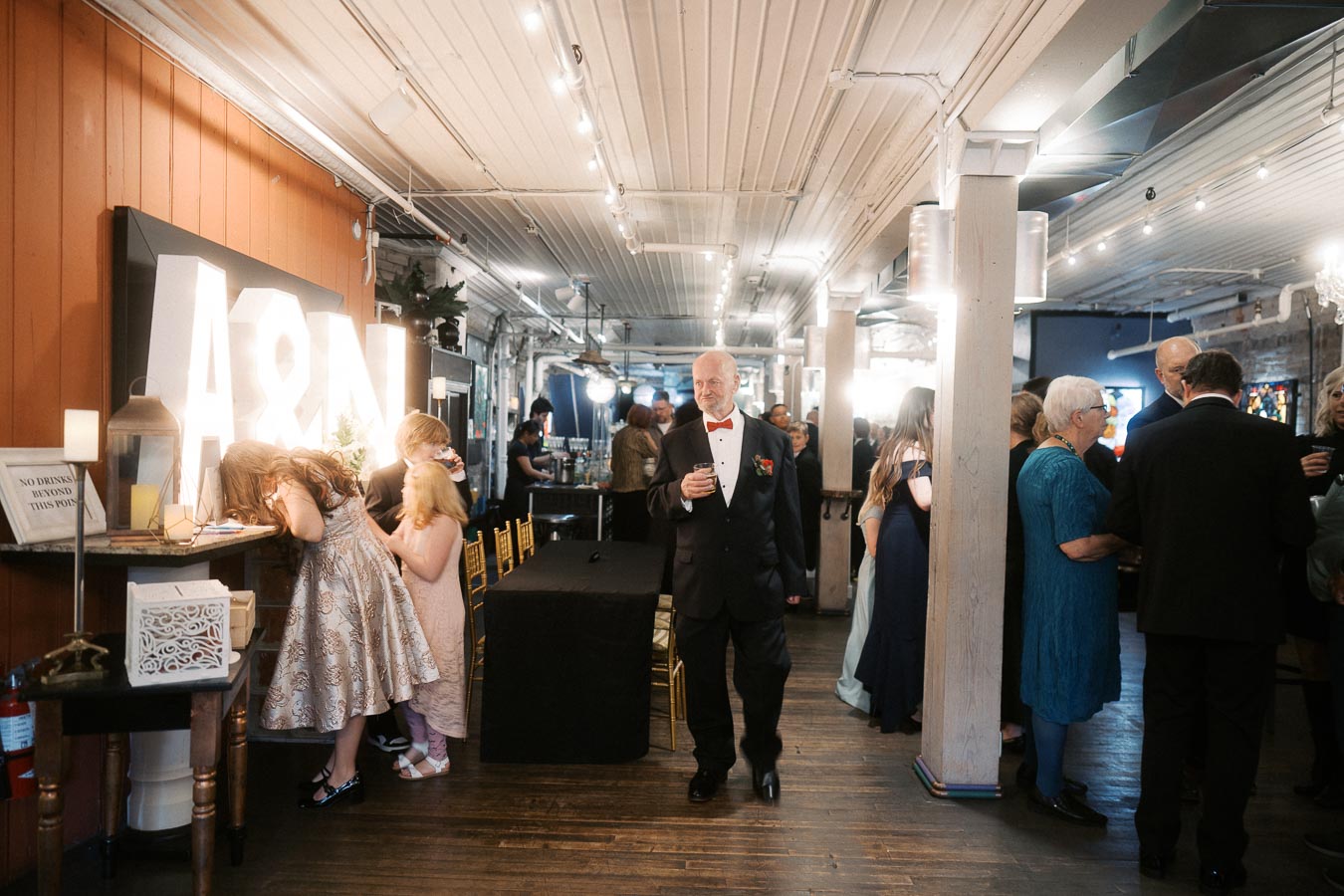 Elegant indoor event celebration with guests mingling and a large illuminated A&N sign in the background. A man in a suit walks toward the camera, holding a drink, while others engage in conversations and children explore.