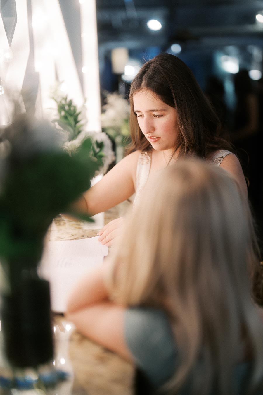 Young girl reading a book at a well-lit marble countertop with decorative plants in the background.