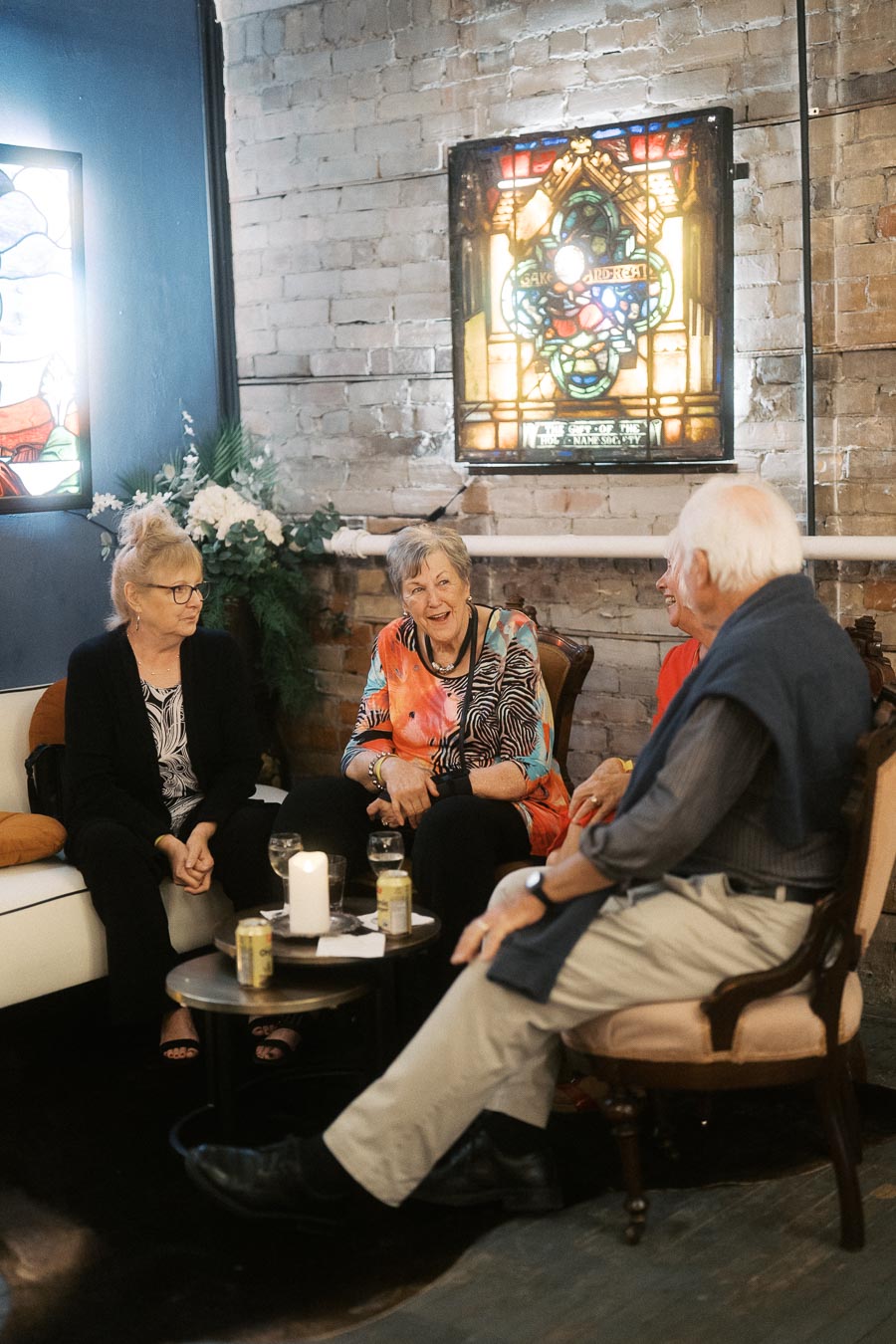 A group of elderly people sitting in a cozy lounge area with drinks on a table, illuminated by stained glass windows and soft lighting, engaging in conversation and smiling warmly.