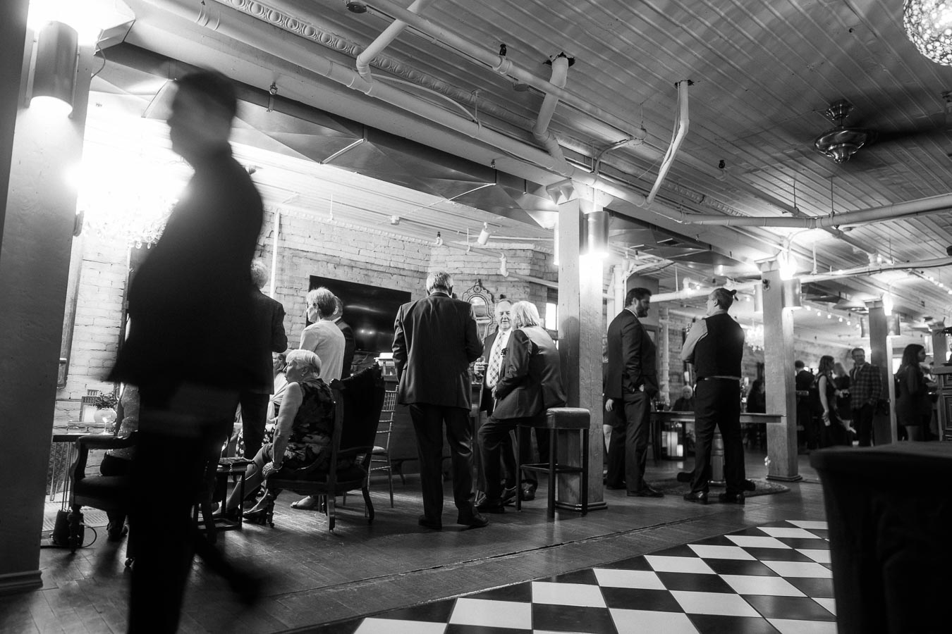 Black and white photo of a lively social event in a stylish venue with people interacting, dressed in elegant attire, with industrial-style decor, and checkered flooring.