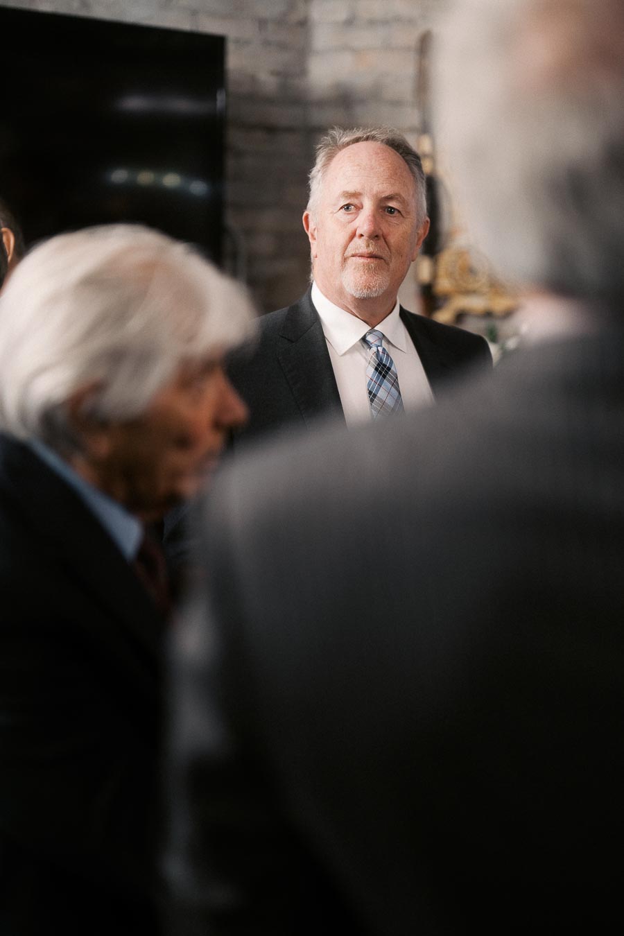 A man in a suit and tie stands attentively in a formal setting, surrounded by others. The environment suggests a professional or social gathering with focus on interpersonal interaction.