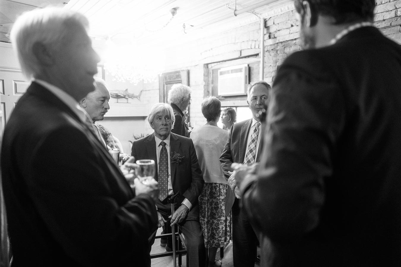 Black and white photo of a group of people in formal attire, socializing in a cozy venue with exposed brick walls. Central figure seated, holding a drink, surrounded by standing guests engaged in conversation.