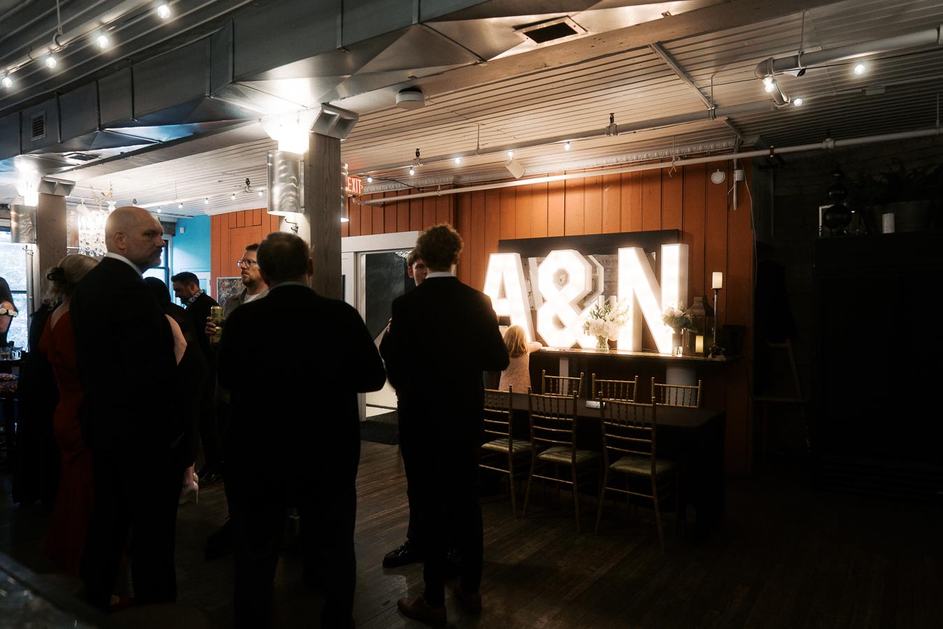 People mingling at an indoor event with a warm atmosphere, featuring large illuminated A & N letters on a wooden backdrop.