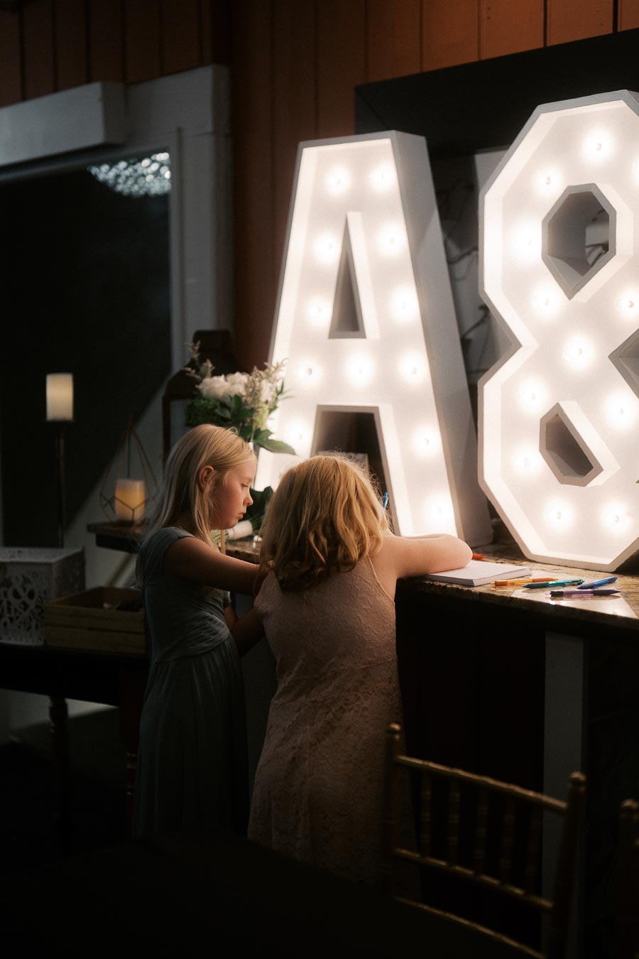 Two children engaged in drawing at a decorated indoor event with illuminated marquee letters A and 8. The warm lighting and decor create a cozy and festive atmosphere.