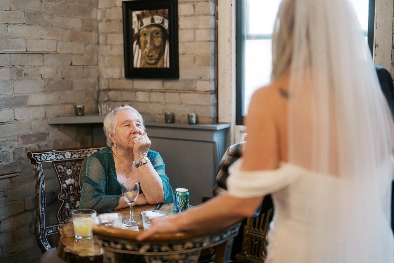 Elderly woman seated at a table with drinks, engaged in conversation with a veiled bride in a rustic brick-walled room, with decorative art on the wall in the background.