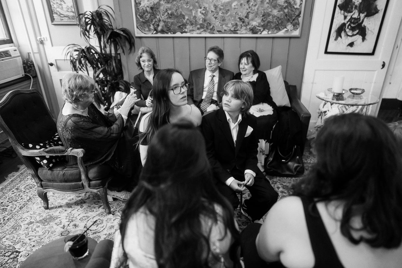 Black and white photo of a group of people seated in a cozy living room, dressed in formal attire, engaged in conversation, surrounded by artwork and plants.