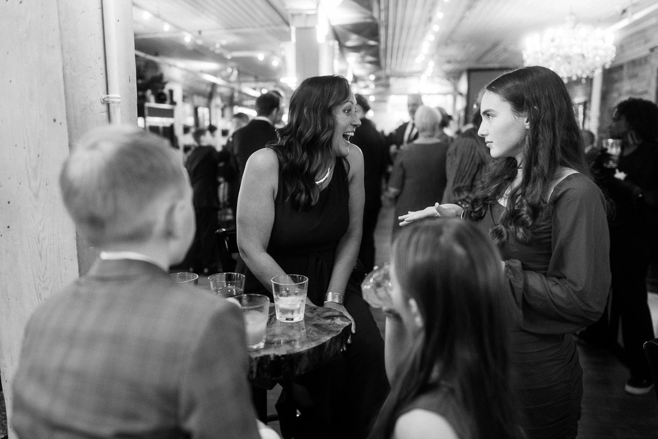Black and white photo of a lively social gathering in a warmly lit venue, featuring a group of people engaging in animated conversation around a small table with drinks. The atmosphere conveys a sense of enjoyment and connection, ideal for event or party-themed content.