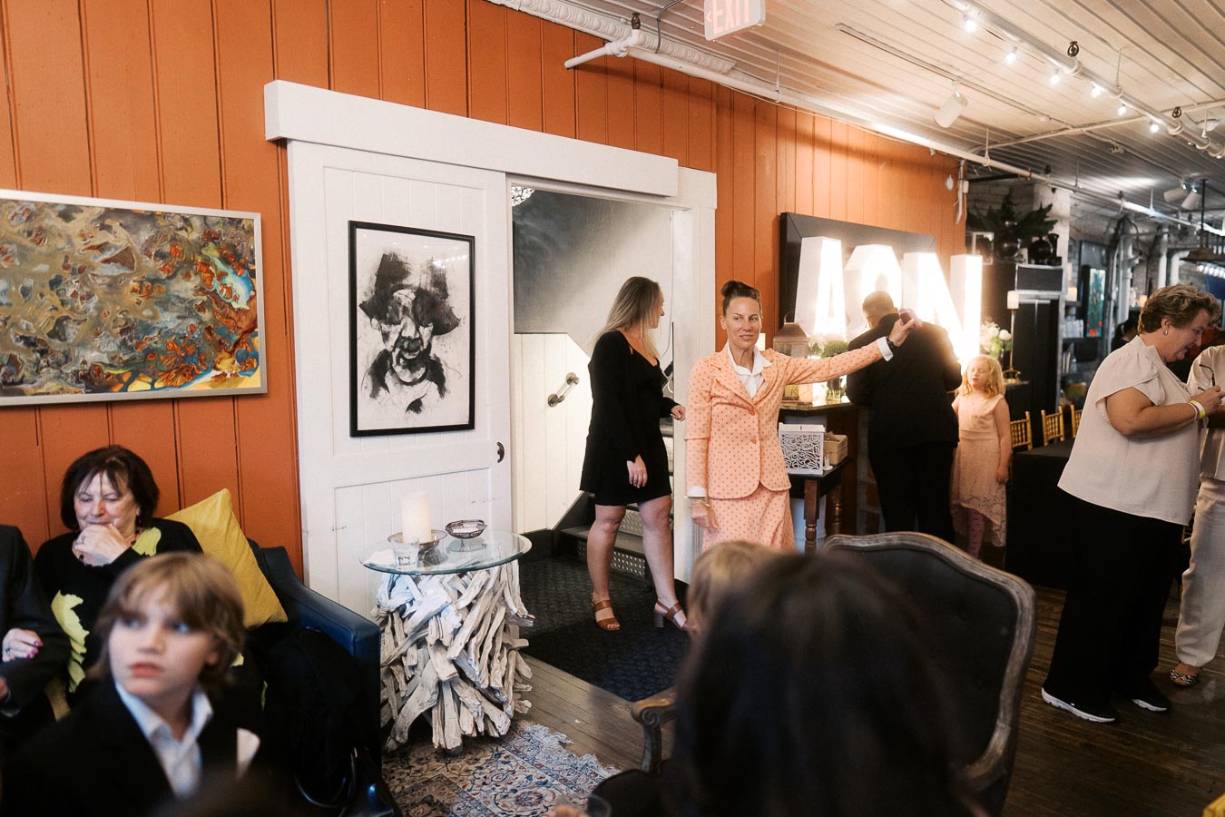 Alt text: Cozy indoor gathering with people socializing in a warmly lit room featuring art on orange walls, a rustic wooden table, and decorative lighting in the background.