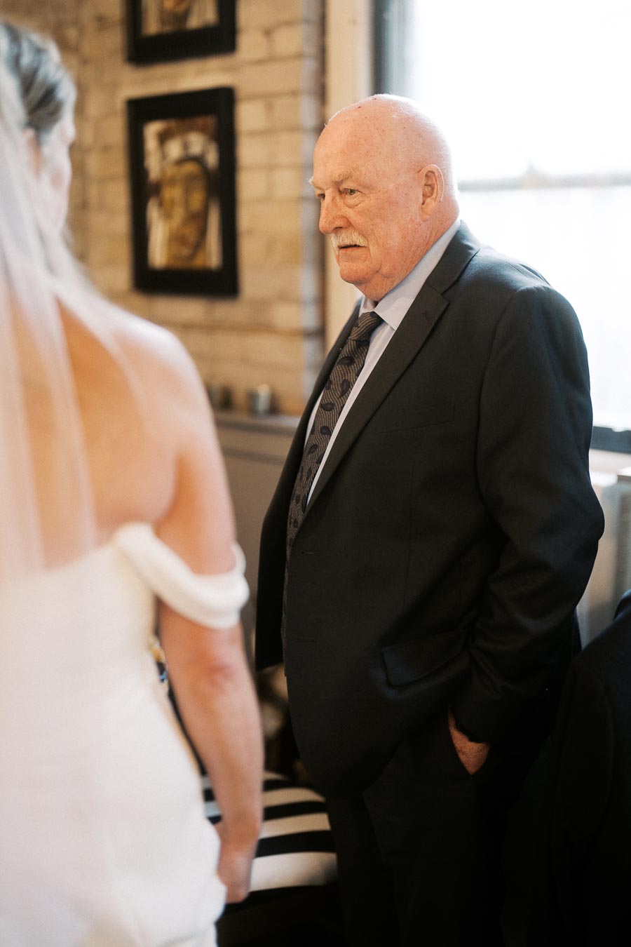 Elderly man in a suit having a conversation with a bride in a wedding dress, indoors with brick walls.