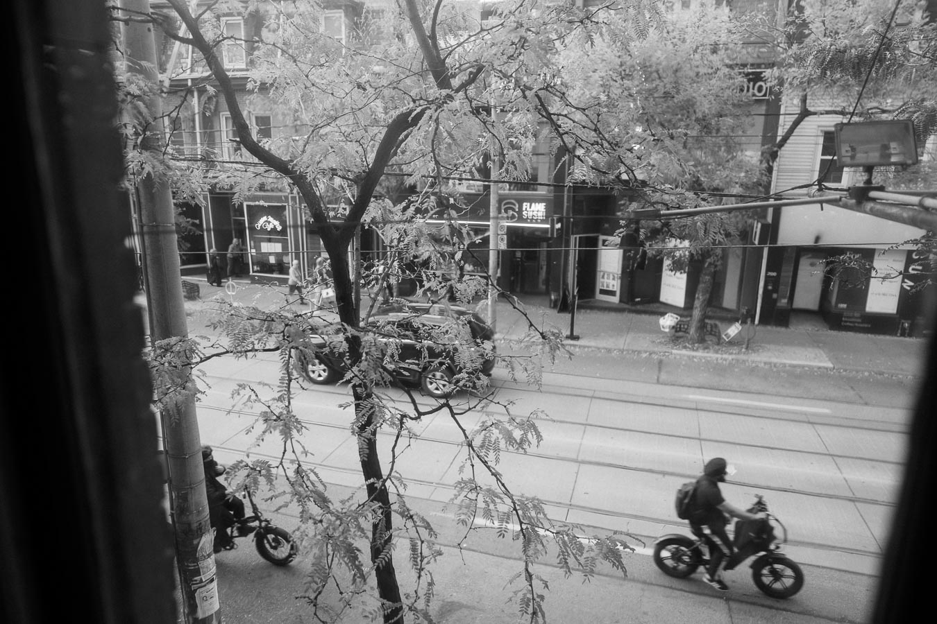 Black and white urban street view featuring two motorcyclists riding along a tree-lined road, with buildings and storefronts, including a sushi restaurant, in the background.