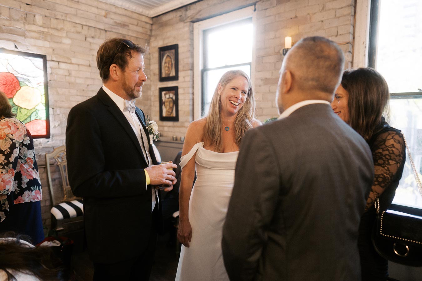 A group of people smiling and conversing at an indoor wedding reception, with a brick wall and colorful stained glass window in the background.