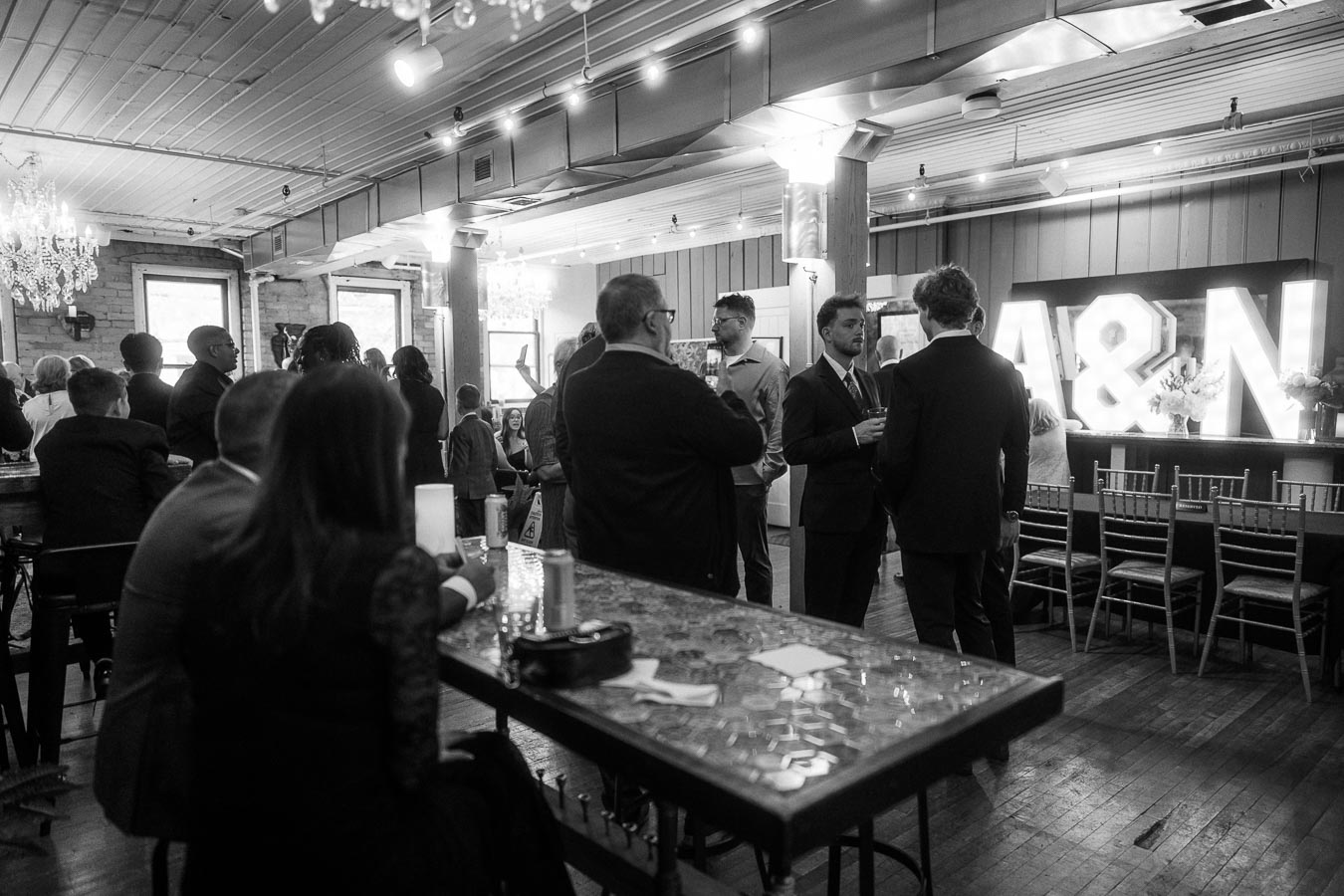 Black and white photo of a social gathering in a chic venue with chandeliers, guests mingling and illuminated letters A&N in the background.