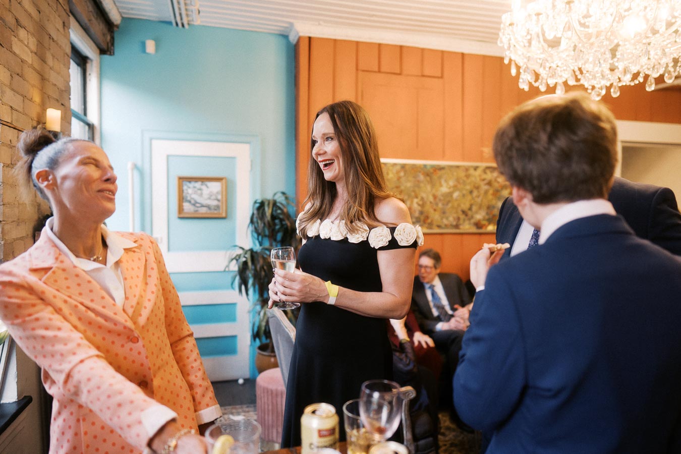 Group of people enjoying a social gathering in a stylish, brightly colored room with modern decor. A woman in a black dress holds a drink while conversing and laughing with other guests. The setting features elegant lighting and a vibrant atmosphere.