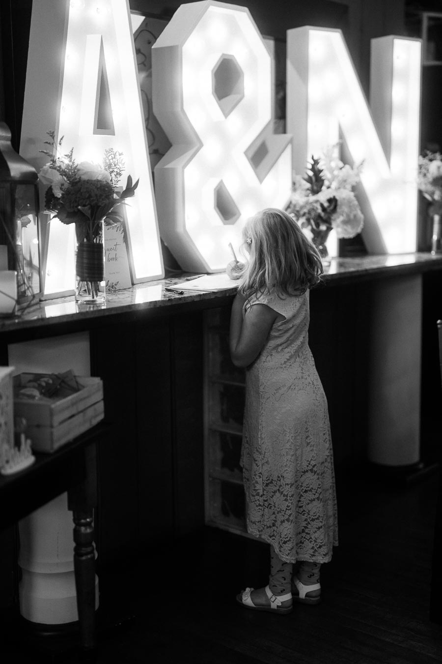 A young girl in a lace dress writing on a surface surrounded by large illuminated letters A & N and flower arrangements in a softly lit room.