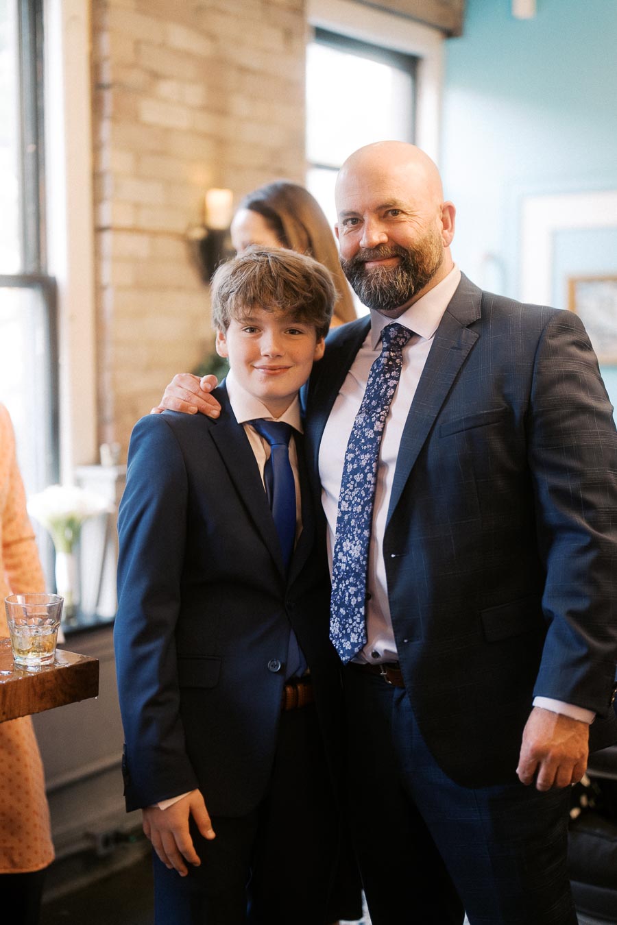 Father and son wearing suits at an indoor social event, standing closely with smiles and arm around each other.