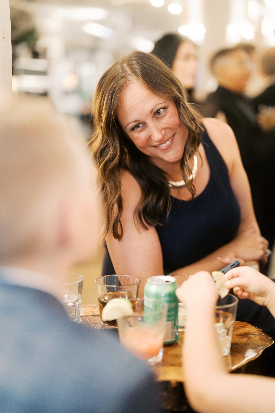 Woman smiling at a social gathering, dressed in a blue dress, sitting at a table with drinks.
