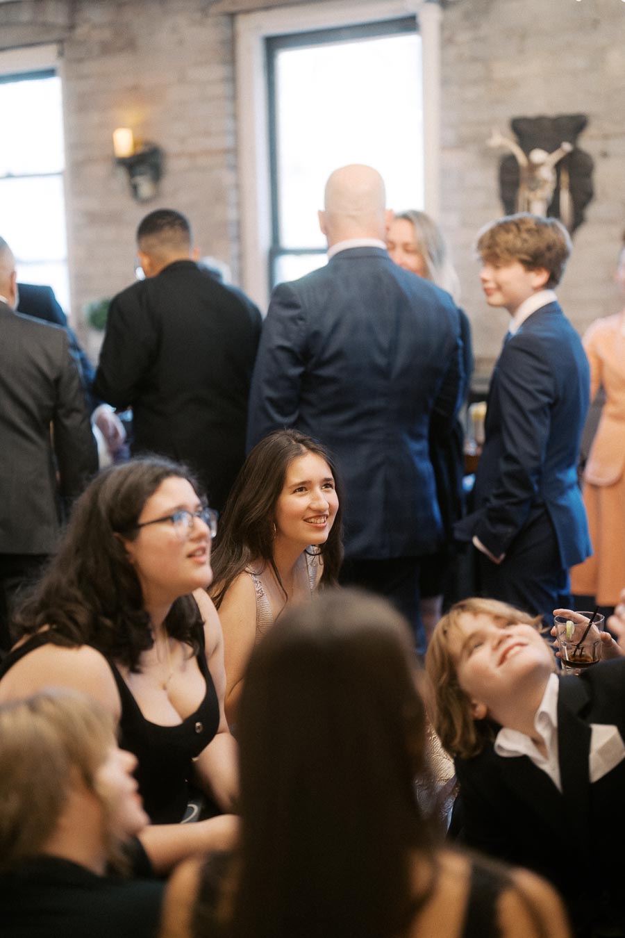A group of people gathered in a stylish indoor setting, with several individuals engaged in conversation and others seated, dressed in formal attire, creating a lively and elegant atmosphere.